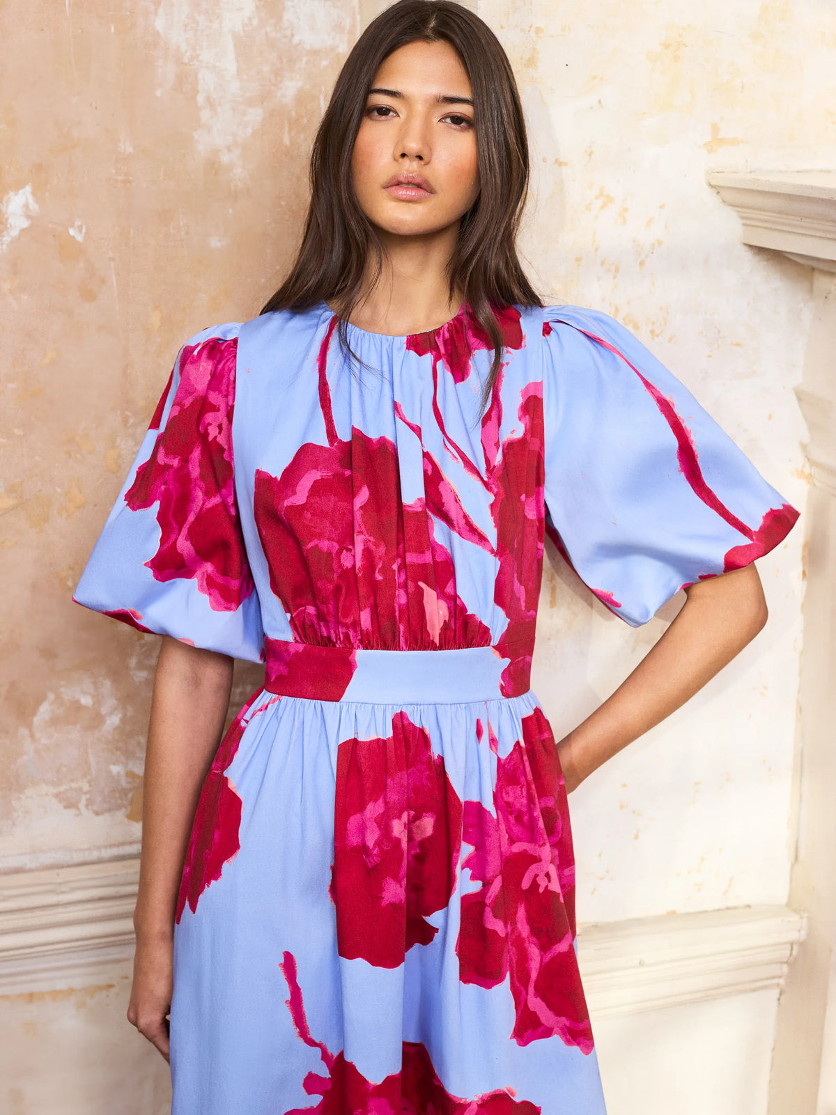 Woman wearing a blue and pink floral dress against a beige wall.