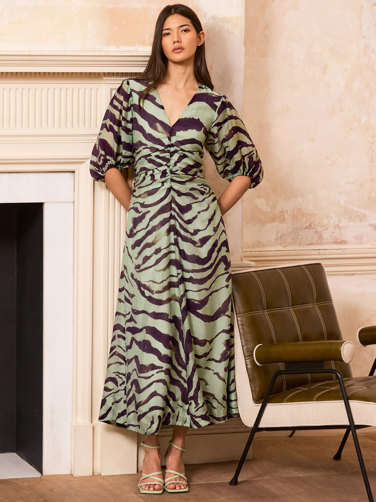Woman wearing a zebra print dress standing in a room with a fireplace and chair.